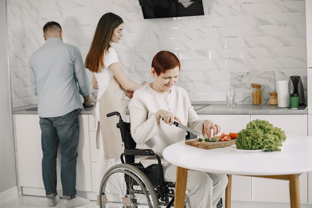 a family prepares dinner in a modern kitchen the children wash dishes while their parent in a wheelchair chops vegetables on the table